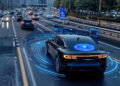 A black car drives on a busy city motorway with digital blue light graphics depicting self-driving or autonomous vehicle technology sensing its surroundings. Other vehicles and buildings are in the background. | FintechZoom