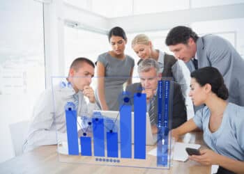 Five business professionals gather around a laptop in a bright office, analyzing data. A transparent screen overlay shows blue bar and line graphs with percentages, indicating a focus on statistics and teamwork. | FintechZoom