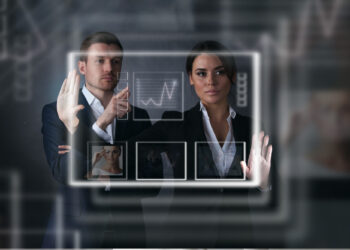Two business professionals stand in front of a virtual transparent screen, interacting with digital charts, graphs, and images, suggesting advanced technology and futuristic data analysis. | FintechZoom