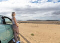 A woman stands beside a light blue car, looking out at a vast desert landscape under a partly cloudy sky with distant mountains on the horizon. | FintechZoom