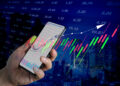 A hand holds a mobile phone displaying a stock market chart, with colourful candlestick graphs and numbers overlaid on a cityscape background, symbolising digital share trading and financial markets. | FintechZoom