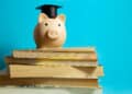 A piggy bank wearing a graduation cap sits on top of a stack of books against a blue background, symbolizing saving money for education. | FintechZoom