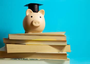 A piggy bank wearing a graduation cap sits on top of a stack of books against a blue background, symbolizing saving money for education. | FintechZoom