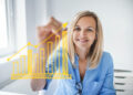 A woman in a blue shirt smiles whilst drawing an upward trending bar and line graph with a yellow marker on a glass surface in a bright office setting. | FintechZoom