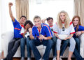 Six young adults wearing football shirts sit on a sofa, some cheering and smiling, one covering their face, holding snacks and drinks, watching a match in a bright living room. | FintechZoom