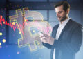 A man in a suit uses his mobile phone whilst standing next to a large glowing Bitcoin symbol with digital financial chart graphics in the background, representing cryptocurrency trading or investment. | FintechZoom