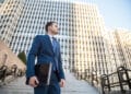 A man in a blue suit holding a laptop stands on outdoor steps in front of a tall, modern office building, looking to the side under a clear sky. | FintechZoom