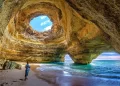 A person stands on the sandy shore inside a large coastal cave with a circular skylight in the ceiling, sunlight streaming in, and turquoise waves entering through wide openings. | FintechZoom