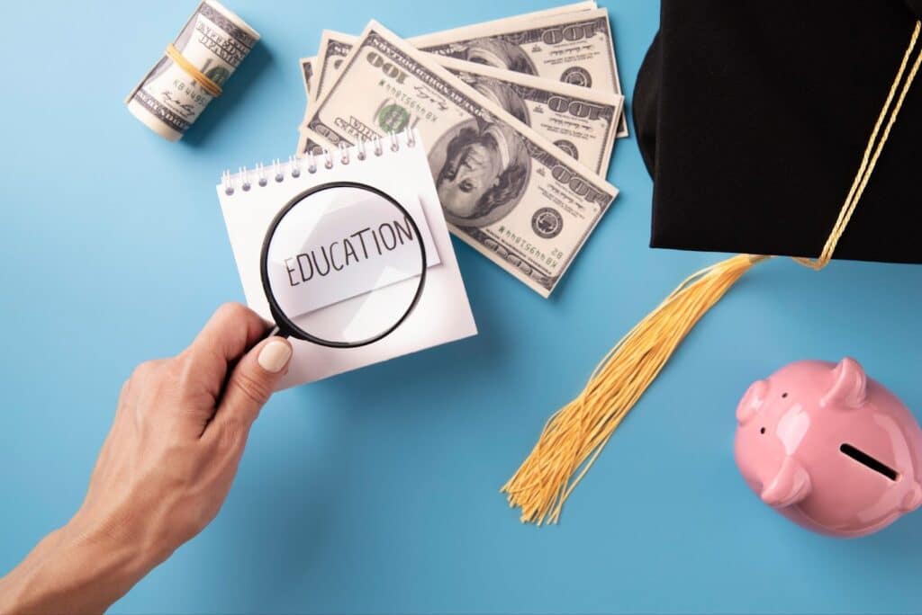 A hand holds a magnifying glass over a notepad with "EDUCATION" written on it. Nearby are US dollar bills, a rolled banknote, a graduation cap, and a pink piggy bank, all on a blue background. | FintechZoom