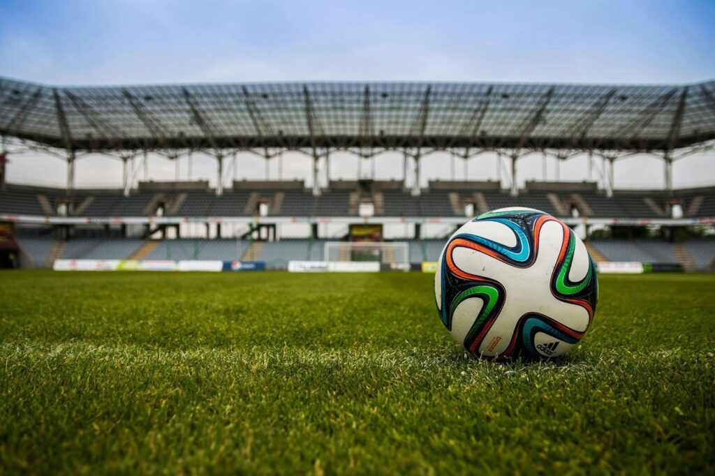 A close-up of a colourful football resting on green grass in an empty stadium, with rows of grey seats and a metal roof visible in the background under a clear sky. | FintechZoom