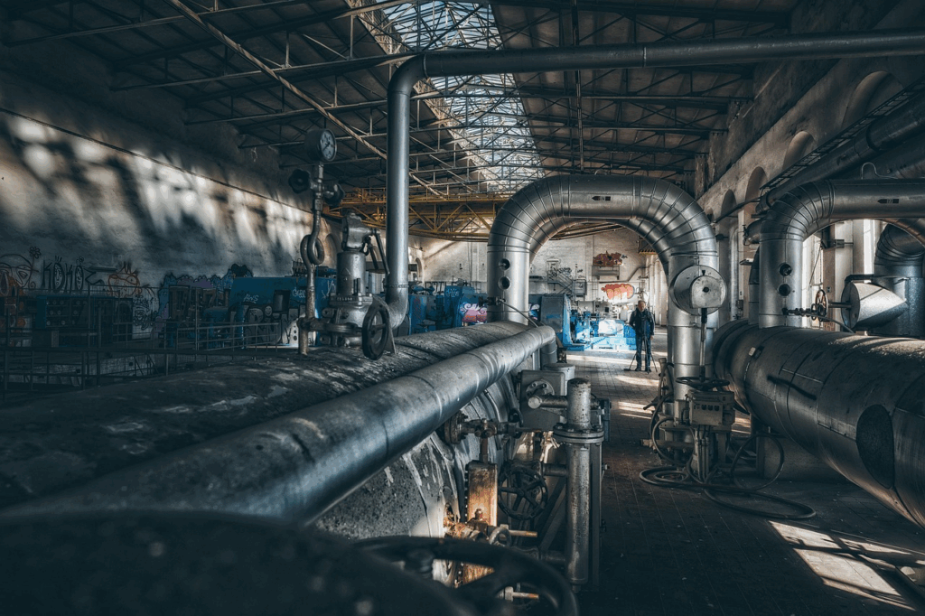 An industrial building with large metal pipes, gauges, and machinery. Sunlight streams through a glass ceiling. A person stands in the background near blue barrels and graffiti-covered walls. | FintechZoom
