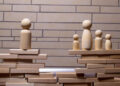 Several wooden figurines stand on stacks of wooden blocks, separated by a gap. One figure is isolated on the left, while four figures stand together on the right, suggesting separation or division. A brick wall is in the background. | FintechZoom