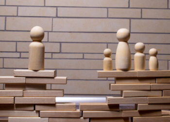 Several wooden figurines stand on stacks of wooden blocks, separated by a gap. One figure is isolated on the left, while four figures stand together on the right, suggesting separation or division. A brick wall is in the background. | FintechZoom
