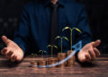 A person in a dark shirt and tie sits behind stacks of coins arranged in ascending order, each topped with a growing plant, symbolising financial growth, with a blue upward arrow overlay. | FintechZoom