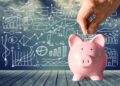 A hand drops a coin into a pink piggy bank on a wooden surface, with various financial charts and graphs drawn on a blackboard in the background. | FintechZoom