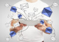 A person holding a tablet with illustrated blue and white envelopes appearing to fly out from the device, representing sending or receiving multiple emails. | FintechZoom