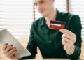 A person in a black shirt holds a red bank card in one hand and a tablet in the other, seated indoors in front of a window with blinds. | FintechZoom