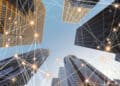 Looking up at tall modern skyscrapers under a blue sky, with glowing connected lines digitally overlaid, representing a network or smart city technology. | FintechZoom