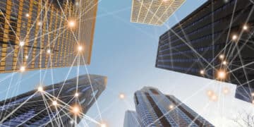 Looking up at tall modern skyscrapers under a blue sky, with glowing connected lines digitally overlaid, representing a network or smart city technology. | FintechZoom