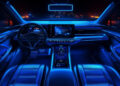 Futuristic car interior with sleek black seats and dashboard, illuminated by vibrant neon blue lights, featuring a large central touchscreen and city lights visible through the windscreen at night. | FintechZoom