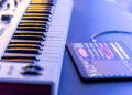 A digital music keyboard sits next to a tablet displaying audio editing software, both on a blue surface with soft lighting. | FintechZoom