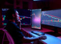 A person sits at a desk with three large monitors displaying colourful share charts and financial data, illuminated by pink and blue lighting, appearing focused and thoughtful. | FintechZoom