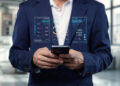 A person in a business suit holds a mobile phone, with digital data charts and graphs projected above the device, representing data analysis and technology in a modern office setting. | FintechZoom