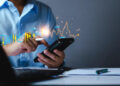 A person in a blue shirt uses a smartphone and stylus at a desk, with digital financial charts and graphs overlaid, representing share market analysis or online trading through a fintech app. | FintechZoom