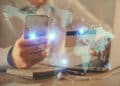 A person holding a mobile phone and a credit card sits at a desk with a laptop, notebook, and pen. A glowing digital world map overlay suggests global online transactions or digital connectivity. | FintechZoom