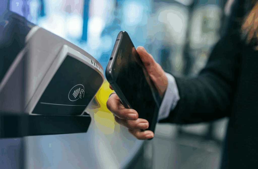 A person holds a mobile phone near a contactless payment terminal, indicating they are making a mobile payment. The background is blurred, focussing on the phone and payment device. | FintechZoom