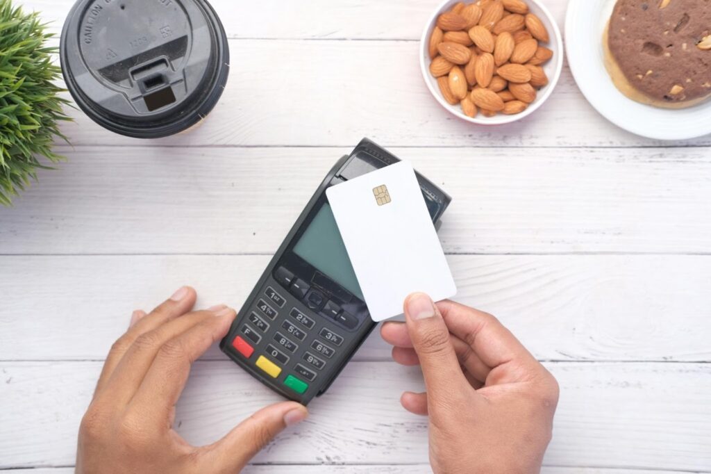 A person holds a white credit card near a payment terminal, preparing for contactless payment. On the table are a coffee cup, a bowl of almonds, a muffin, and a small plant. | FintechZoom
