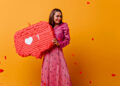 A smiling woman in a pink blouse and long pink skirt holds a large red piñata shaped like a social media like notification with a heart and the number 1. Red confetti hearts are scattered against a yellow background. | FintechZoom