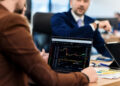 Two people in an office discuss financial data; one holds a laptop displaying share charts and graphs, while the other gestures in conversation. Papers and a tablet are also on the desk. | FintechZoom