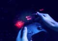 A person holds a game controller with a futuristic digital interface and glowing red and blue graphics overlaying the scene, representing technology and gaming concepts. | FintechZoom