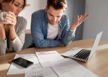 A worried couple sits at a table, looking at bills and documents spread out next to a laptop and a mobile. One person holds a mug, while the other gestures with frustration. Sunlight streams in from the side. | FintechZoom