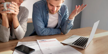 A worried couple sits at a table, looking at bills and documents spread out next to a laptop and a mobile. One person holds a mug, while the other gestures with frustration. Sunlight streams in from the side. | FintechZoom