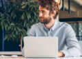 A man with brown hair and a beard sits at a desk, looking to the side, with a laptop open in front of him and a large leafy plant in the background. | FintechZoom