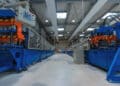 Wide-angle view of an industrial factory interior with large blue and orange automated machines lining both sides of a clean, spacious aisle under a high, corrugated metal ceiling with bright overhead lights. | FintechZoom