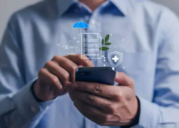 A person in a blue shirt uses a mobile phone, with insurance-related icons such as a document, umbrella, shield, and leaf digitally illustrated above the device, symbolising digital insurance services. | FintechZoom