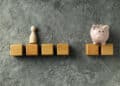 A row of wooden blocks on a grey textured surface, with a wooden figure standing on one block and a pink piggy bank on another, separated by a gap. | FintechZoom