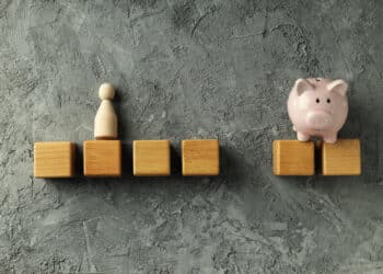 A row of wooden blocks on a grey textured surface, with a wooden figure standing on one block and a pink piggy bank on another, separated by a gap. | FintechZoom