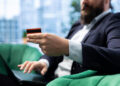 A person in a suit sits on a green chair using a laptop, holding a credit card in one hand. The background is blurred, suggesting an office or modern indoor setting. | FintechZoom