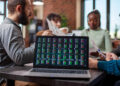 A laptop displaying stock market data sits on a table in the foreground, whilst four people sit around the table in the background, discussing documents in a modern office setting. | FintechZoom