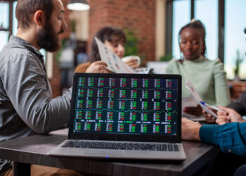 A laptop displaying stock market data sits on a table in the foreground, whilst four people sit around the table in the background, discussing documents in a modern office setting. | FintechZoom