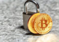 Two gold-coloured Bitcoin coins leaning against a silver padlock on a textured grey surface, symbolising cryptocurrency security or protection. | FintechZoom