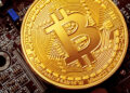 A close-up of a gold-coloured physical Bitcoin coin displaying the Bitcoin symbol, placed on top of a computer motherboard with visible electronic components. | FintechZoom