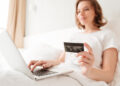 A woman sits on a bed using a laptop whilst holding a credit card, appearing to shop or make a payment online. She is dressed in white and the room is softly lit. | FintechZoom