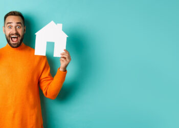 A smiling man in an orange jumper holds a stack of dollar notes in one hand and a white paper cut-out of a house in the other, standing against a teal background with space on the right. | FintechZoom