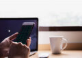 A person holds a mobile phone displaying share market data in front of a laptop and a white coffee cup on a wooden desk near a window. | FintechZoom