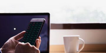 A person holds a mobile phone displaying share market data in front of a laptop and a white coffee cup on a wooden desk near a window. | FintechZoom